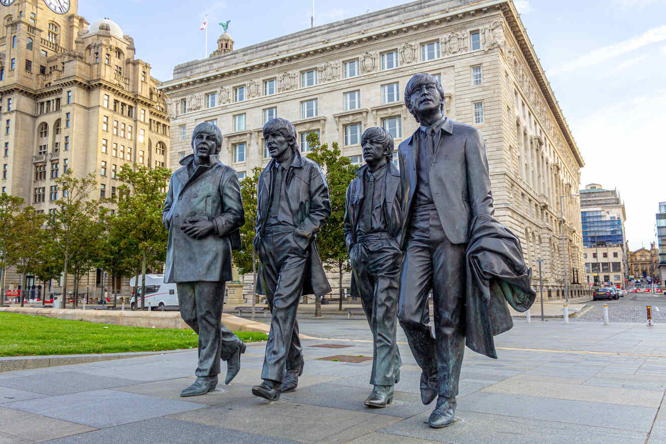 Bronze statues of The Beatles stride across Liverpool's waterfront, set against the backdrop of the iconic Liver Building, under a clear sky