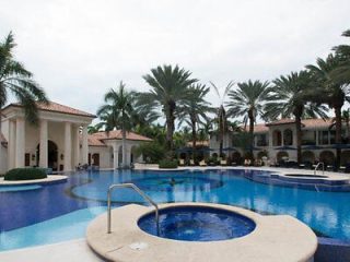 A lavish resort pool area with blue waters, surrounded by palm trees and a classical style building with arches and columns.