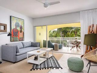 A modern living room opening onto a balcony with tropical views, featuring a gray couch, a white coffee table, and vibrant artwork on the wall.