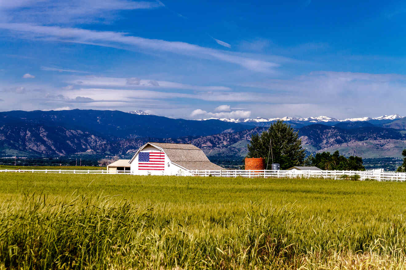 A white barn with an American flag painted on it stands in a green field with mountains in the background under a clear blue sky.