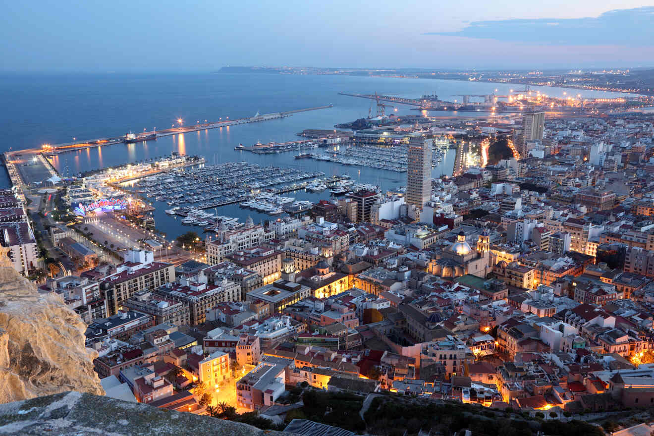 Aerial view of a coastal city at dusk, featuring a marina with numerous boats, tall buildings, and a mix of residential and commercial areas illuminated by city lights.