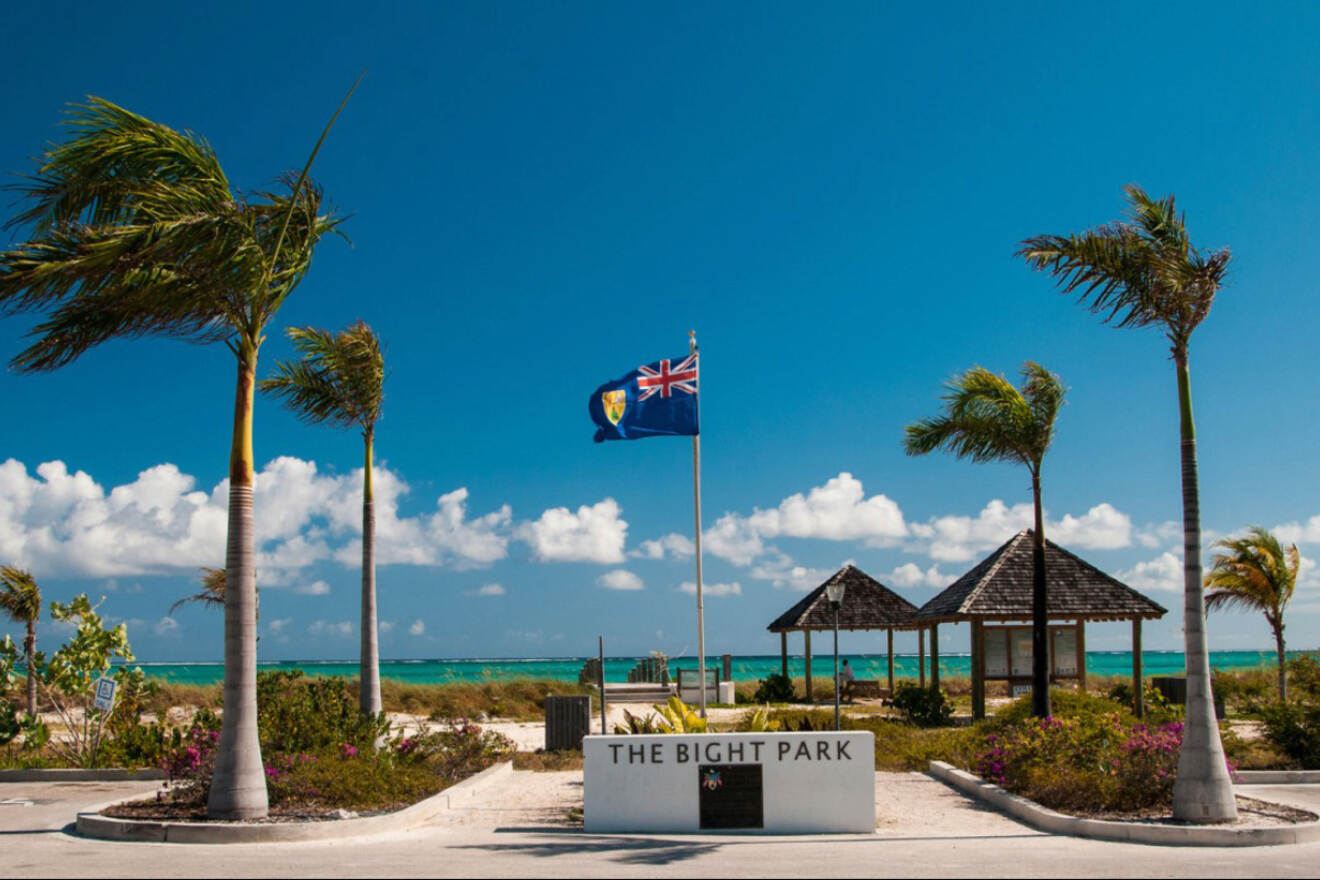 The entrance to The Bight Park with the national flag of the Turks and Caicos Islands waving above, palm trees and ocean in the distance under a blue sky with scattered clouds.