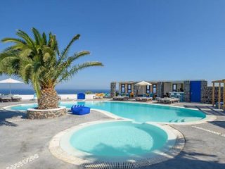 Luxurious resort pool with a palm tree on a bright day in Mykonos, overlooking the serene blue ocean.