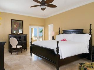 An elegant bedroom with a dark wood four-poster bed and white bedding, a classic writing desk, and balcony doors opening to a tropical view.