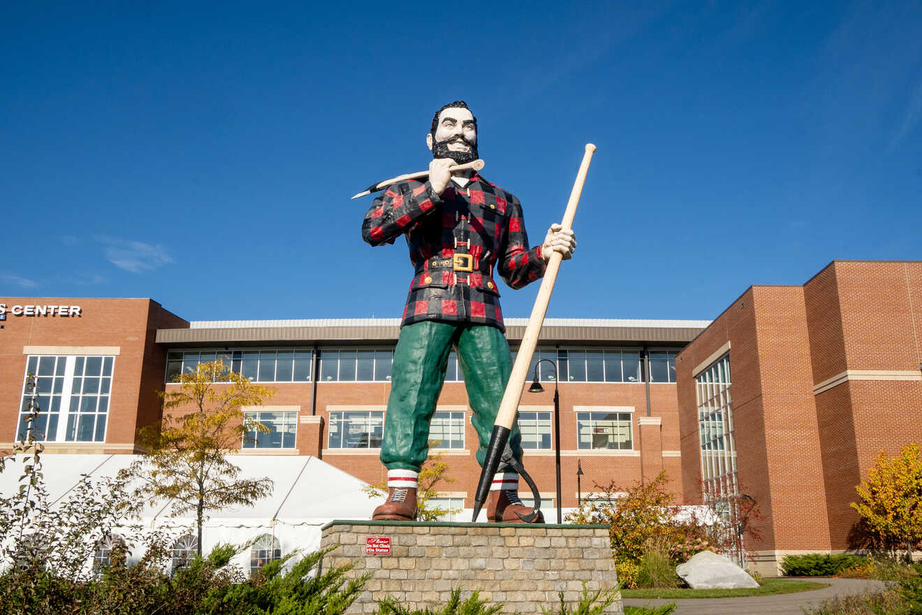 Giant statue of a lumberjack holding an axe, symbolizing the local heritage in front of a modern building with autumn foliage.
