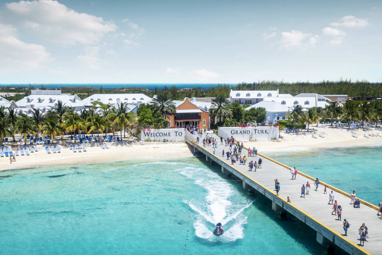 A welcoming view of Grand Turk with a "Welcome to Grand Turk" sign, tourists walking on the pier, and a turquoise ocean surrounding the sandy beach.