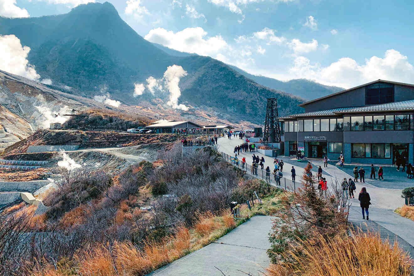 Tourists visiting a geothermal site with steam vents and walking paths, with a modern visitor center and mountains in the background
