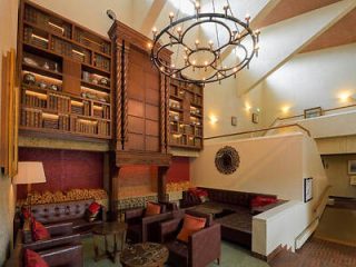 Luxurious lodge interior with high ceiling, chandelier, and a grand bookshelf, furnished with plush red and brown sofas and a central fireplace