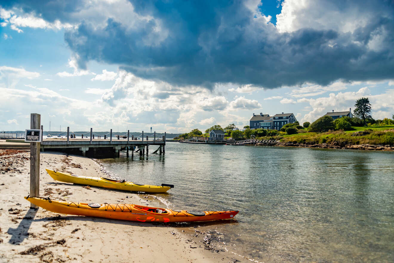 Two yellow kayaks resting on the sandy shore of a peaceful beach with a pier and homes in the background.
