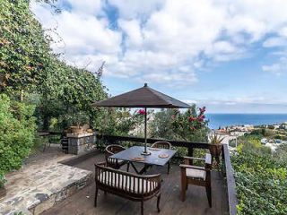 Rustic terrace with wooden furniture and a parasol overlooking the sea and a lush garden.
