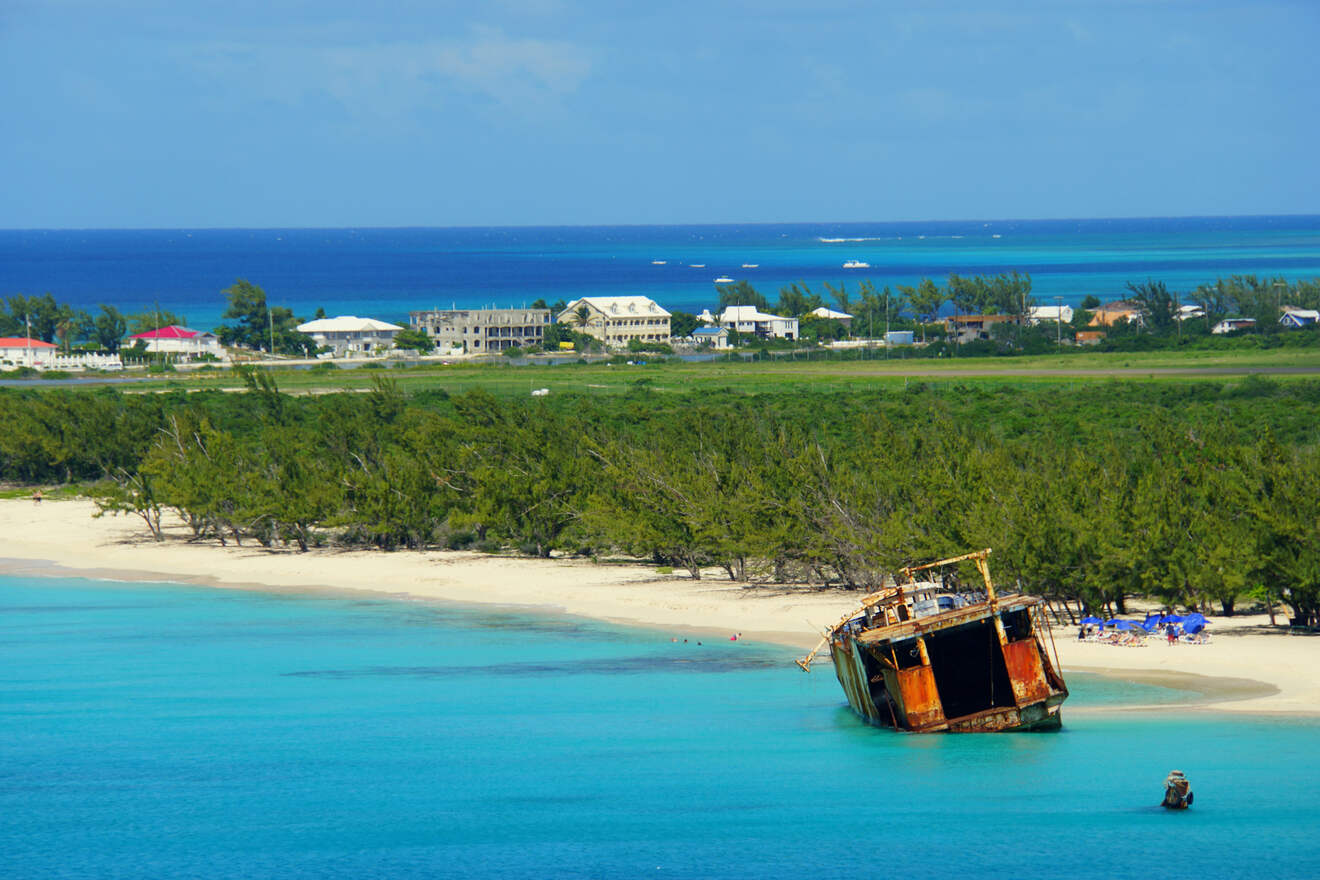 A shipwreck on a sandy beach at the edge of turquoise waters, with a backdrop of tropical trees and distant buildings under a clear blue sky.