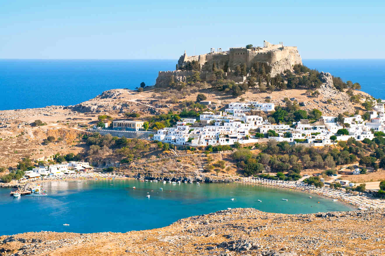 Medieval fortress on a hill overlooking the white buildings of Lindos village and a serene blue bay in Rhodes, Greece.
