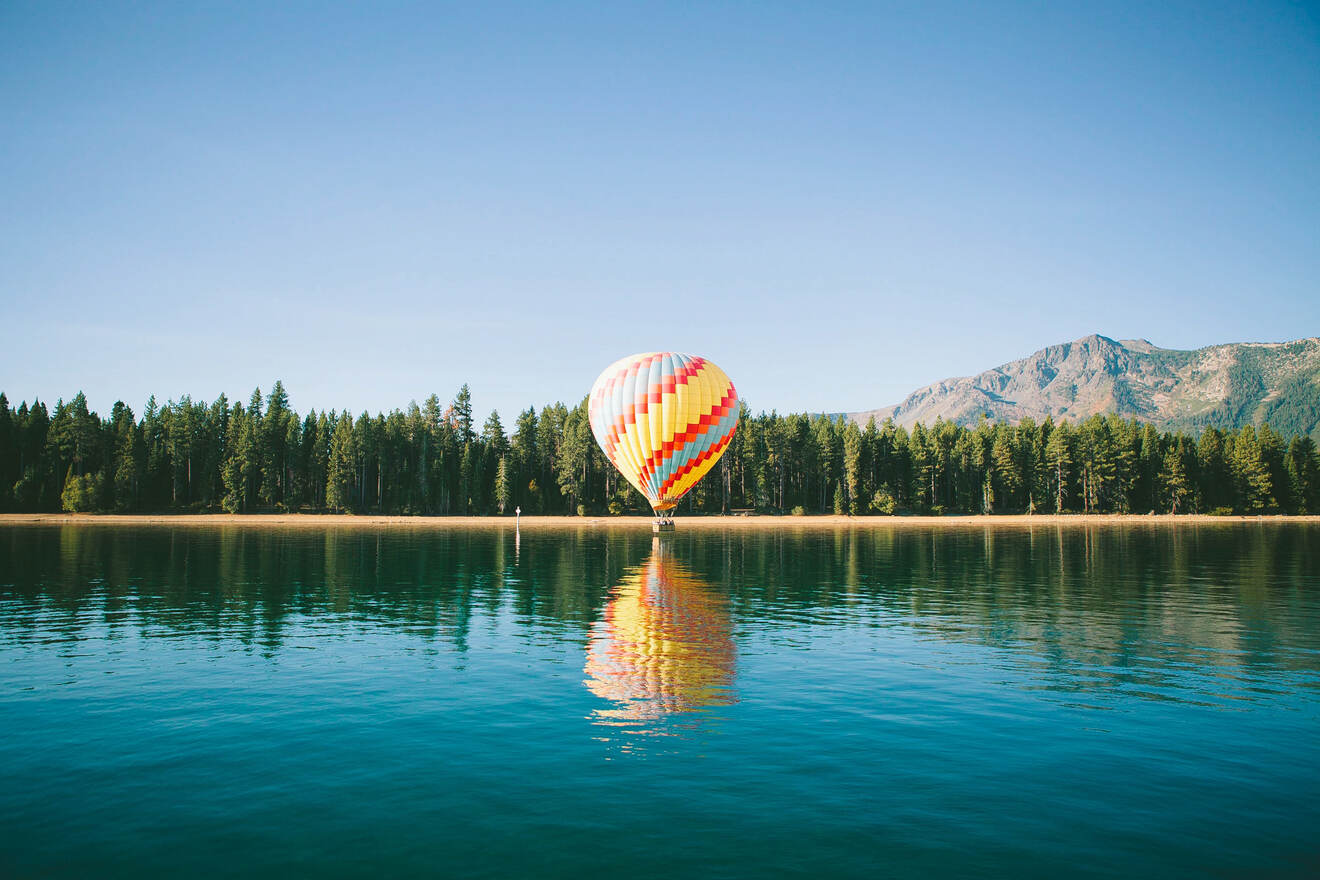 Colorful hot air balloon reflecting on the calm waters of a lake surrounded by a forest and mountains in the background.
