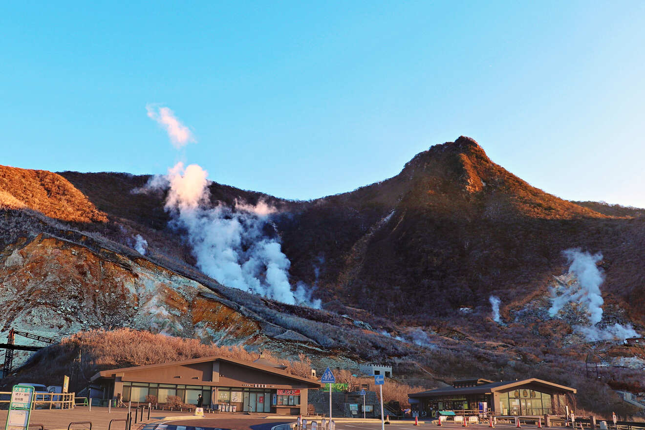Geothermal valley at dusk with steam vents, a walking path, and a visitor center, with a backdrop of mountains under a gradient sky