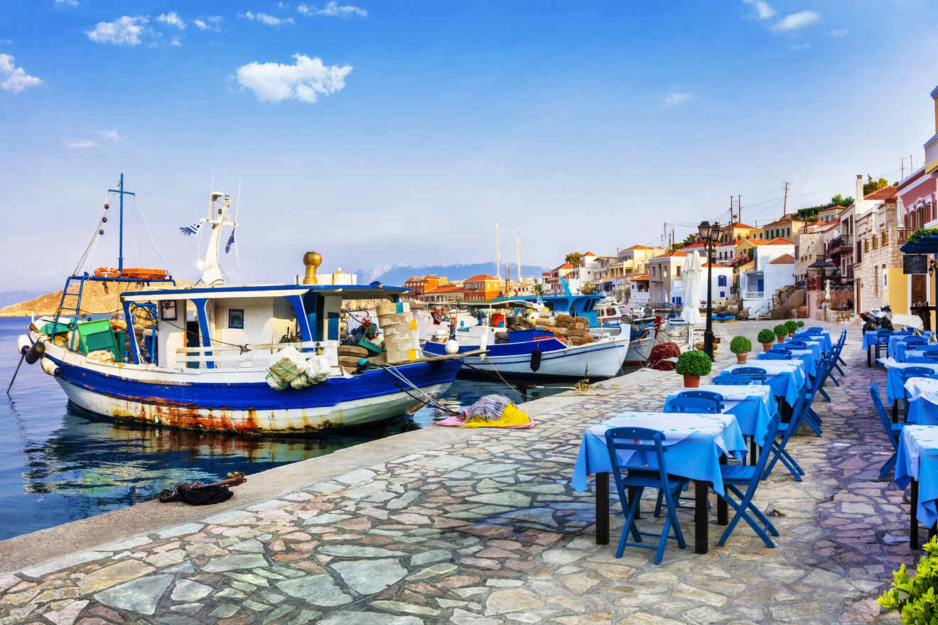 Quaint harbor scene with traditional Greek fishing boats moored alongside a seafront lined with blue tables and chairs.
