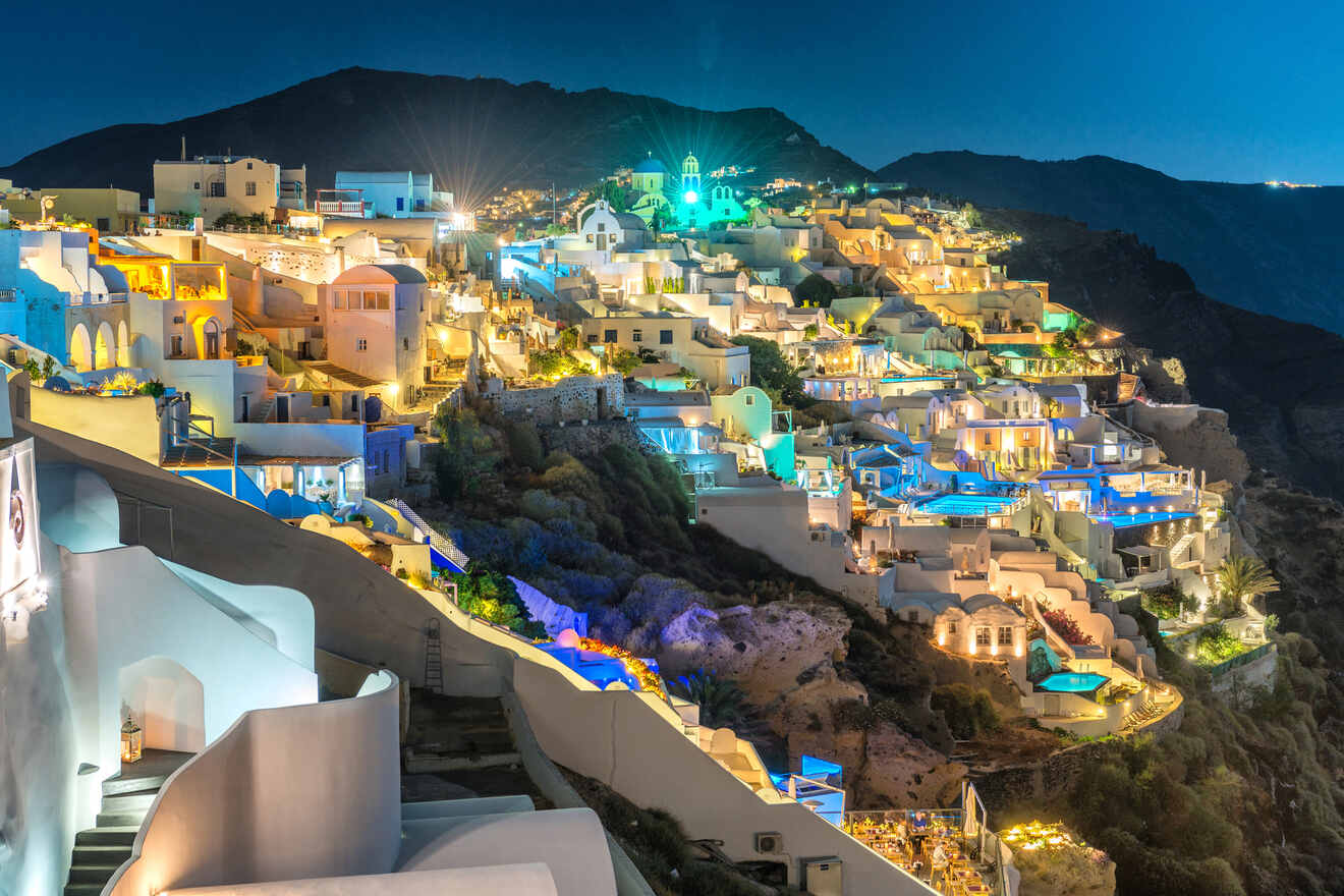 Nighttime view of the illuminated hillside town of Oia in Santorini, with houses and pools glowing under the starry sky.
