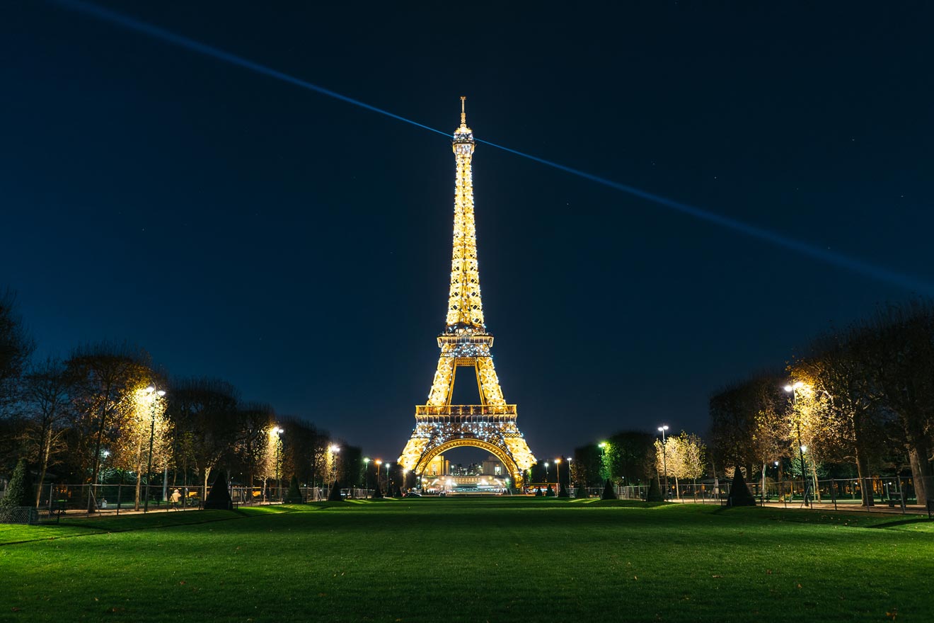 The Eiffel Tower in Paris, illuminated against a dark night sky with a beam of light shining from its top, viewed from a grassy park.