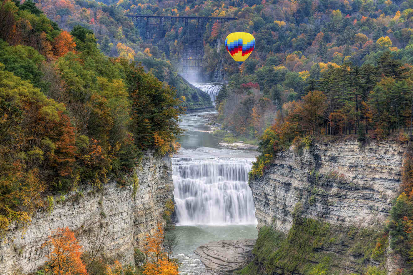 A vibrant hot air balloon floats over Letchworth State Park with autumn-colored foliage, a waterfall, and the historic Portageville Bridge in the backdrop.