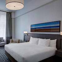 A modern hotel bedroom with a large bed, plush headboard, and a seating area by the window.