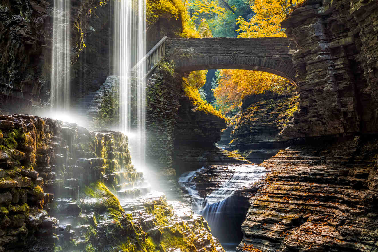 A stunning view of Watkins Glen State Park where cascading waterfalls are framed by a stone bridge and sunlit golden-hued autumn leaves