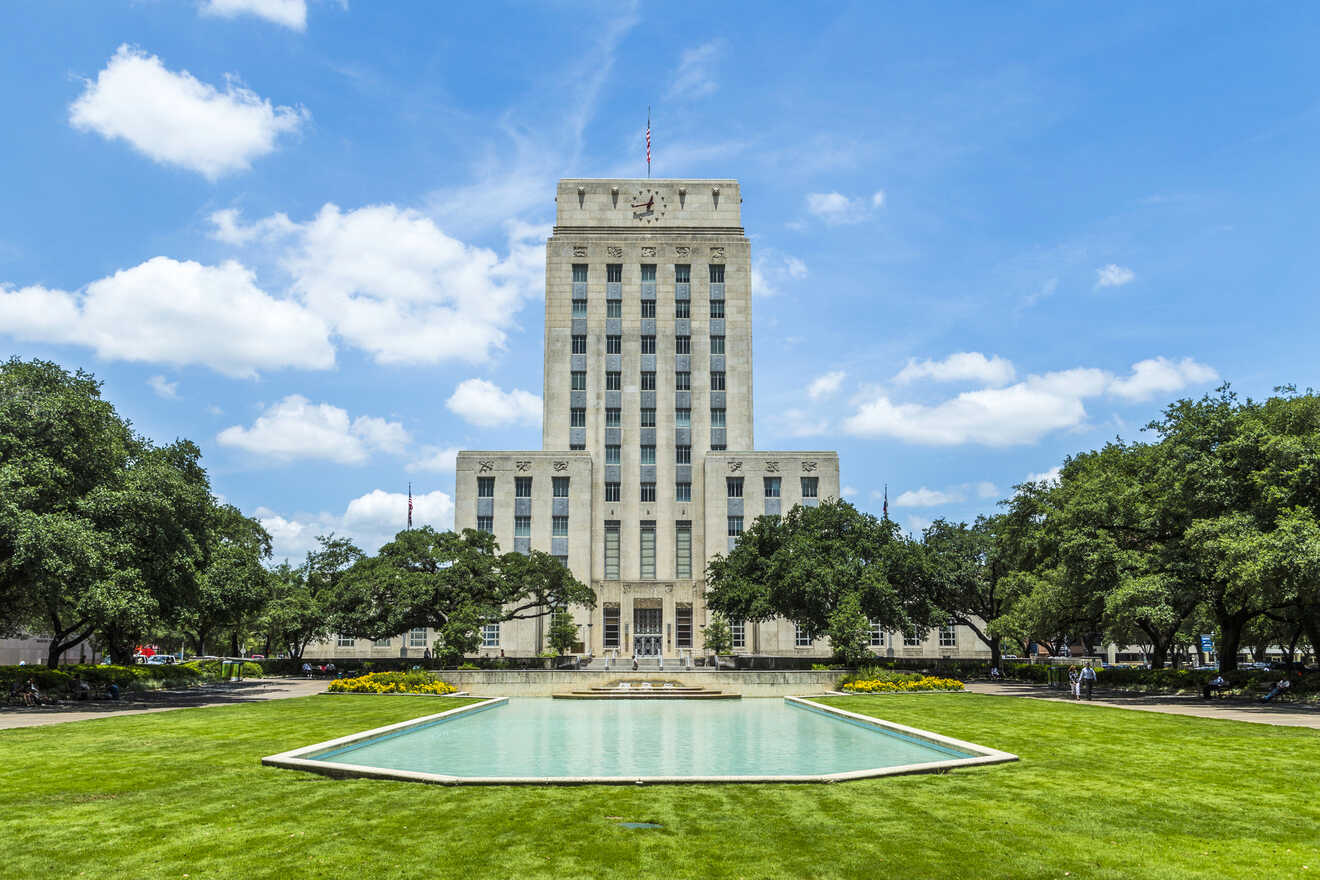 A tall, rectangular building stands behind a rectangular reflecting pool with green grass and trees surrounding it under a blue sky with scattered clouds.