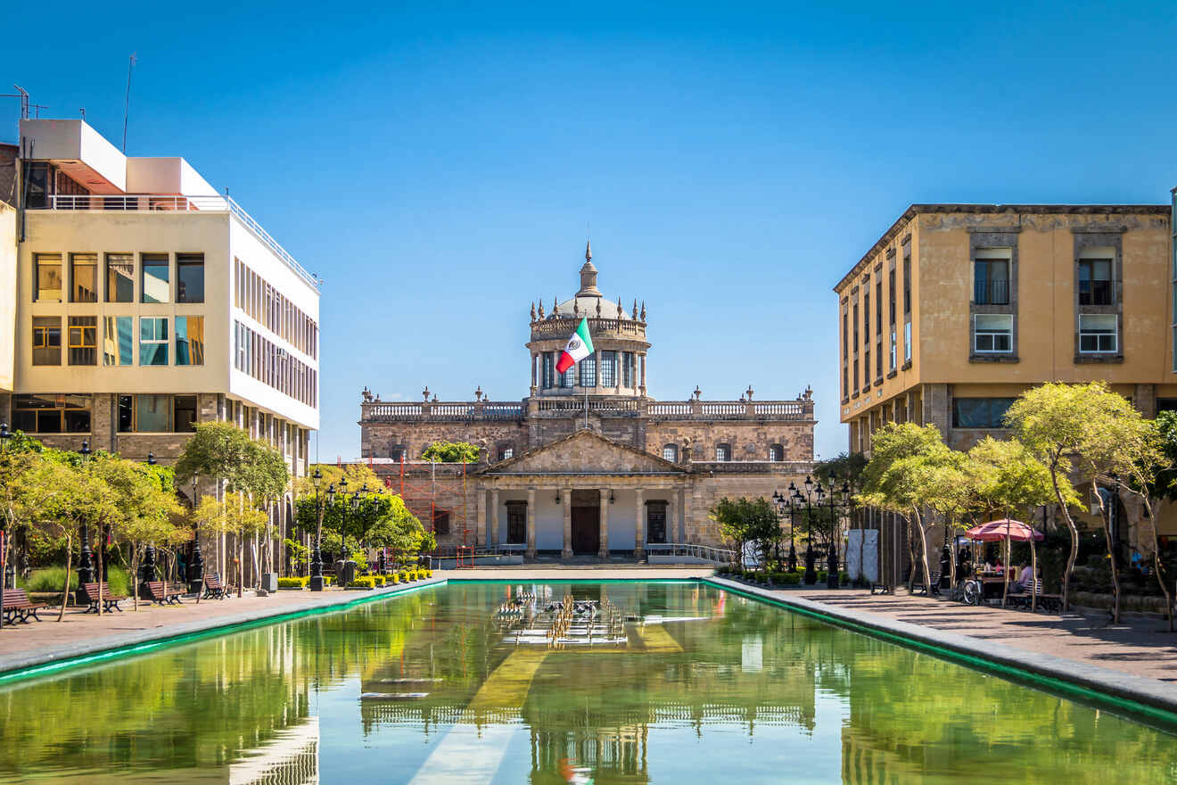 Scenic view of Guadalajara's Hospicio Cabañas with a Mexican flag atop, reflected in a long rectangular fountain, flanked by modern and historic architecture
