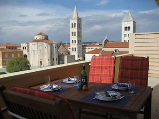 A rooftop dining area with a set table and red checkered chairs, offering a view of historic stone churches and bell towers in the background.