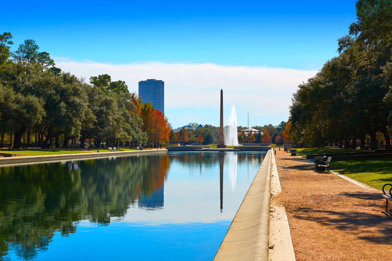 A tree-lined reflecting pool with a tall fountain and an obelisk in a city park under a clear blue sky.