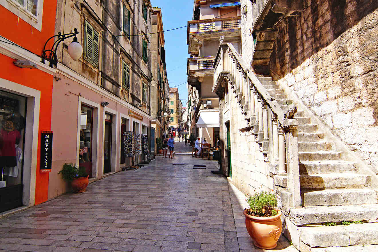 A narrow cobblestone street lined with shops and old buildings, with an outdoor staircase on the right.
