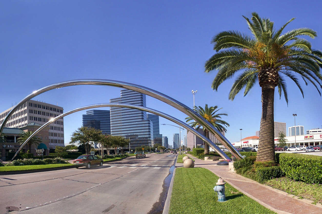 A city street lined with palm trees and tall buildings. Two large, curved metal arches span across the center of the road.