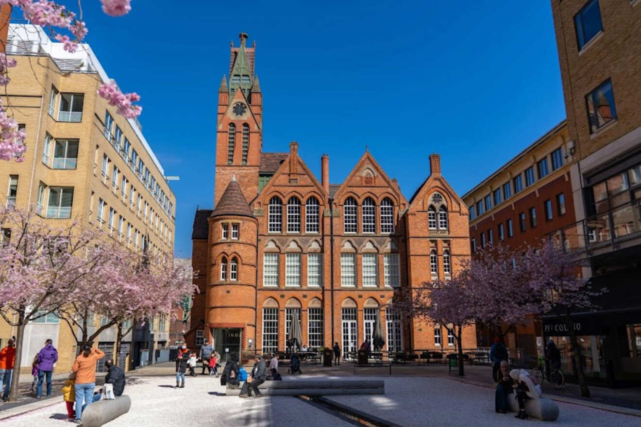 Urban plaza with people enjoying a sunny day, surrounded by flowering trees and historical red brick architecture with tall pointed towers