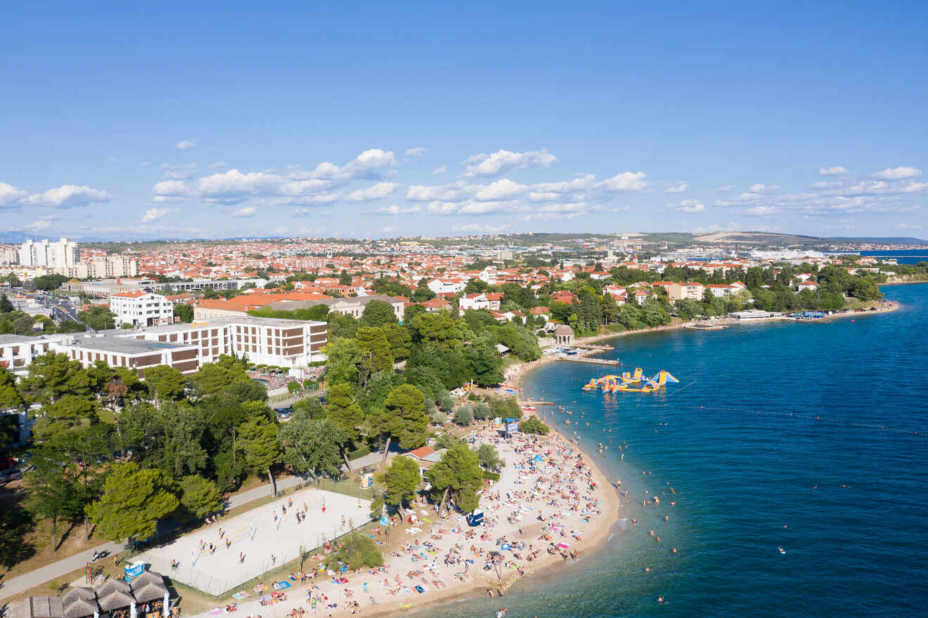 A panoramic view of a beach crowded with people, adjacent to a city with red-roofed buildings and green trees