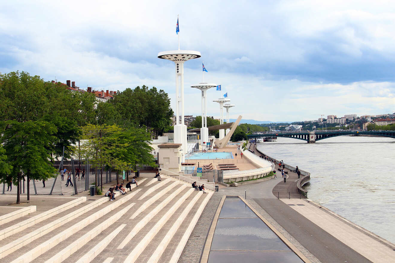 Riverside promenade in Lyon with distinctive futuristic white structures and blue flags, overlooking the Rhône River, with people relaxing along the steps