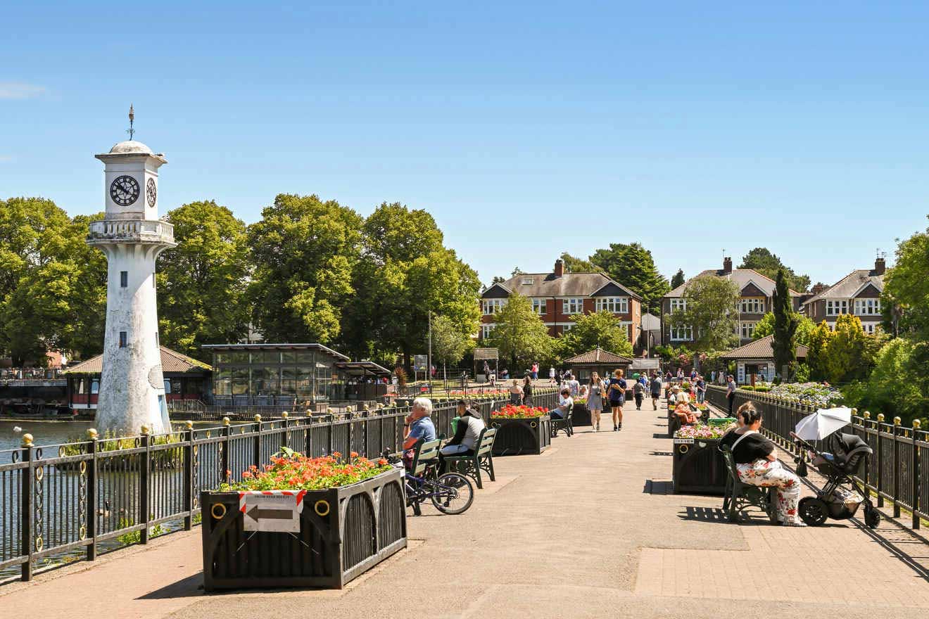 A busy riverside walkway with a lighthouse clock tower, blooming flower beds, and people enjoying the sunny weather