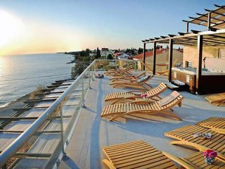 A rooftop terrace with wooden lounge chairs, a hot tub, and a scenic view of the sea at sunset.