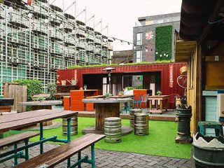 Outdoor urban eatery with vibrant red container bar, green artificial turf, and wooden picnic tables, surrounded by industrial scaffolding
