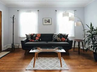A cozy and well-lit living room with a sleek black couch adorned with patterned throw pillows, a stylish arched floor lamp, and two large windows allowing natural light