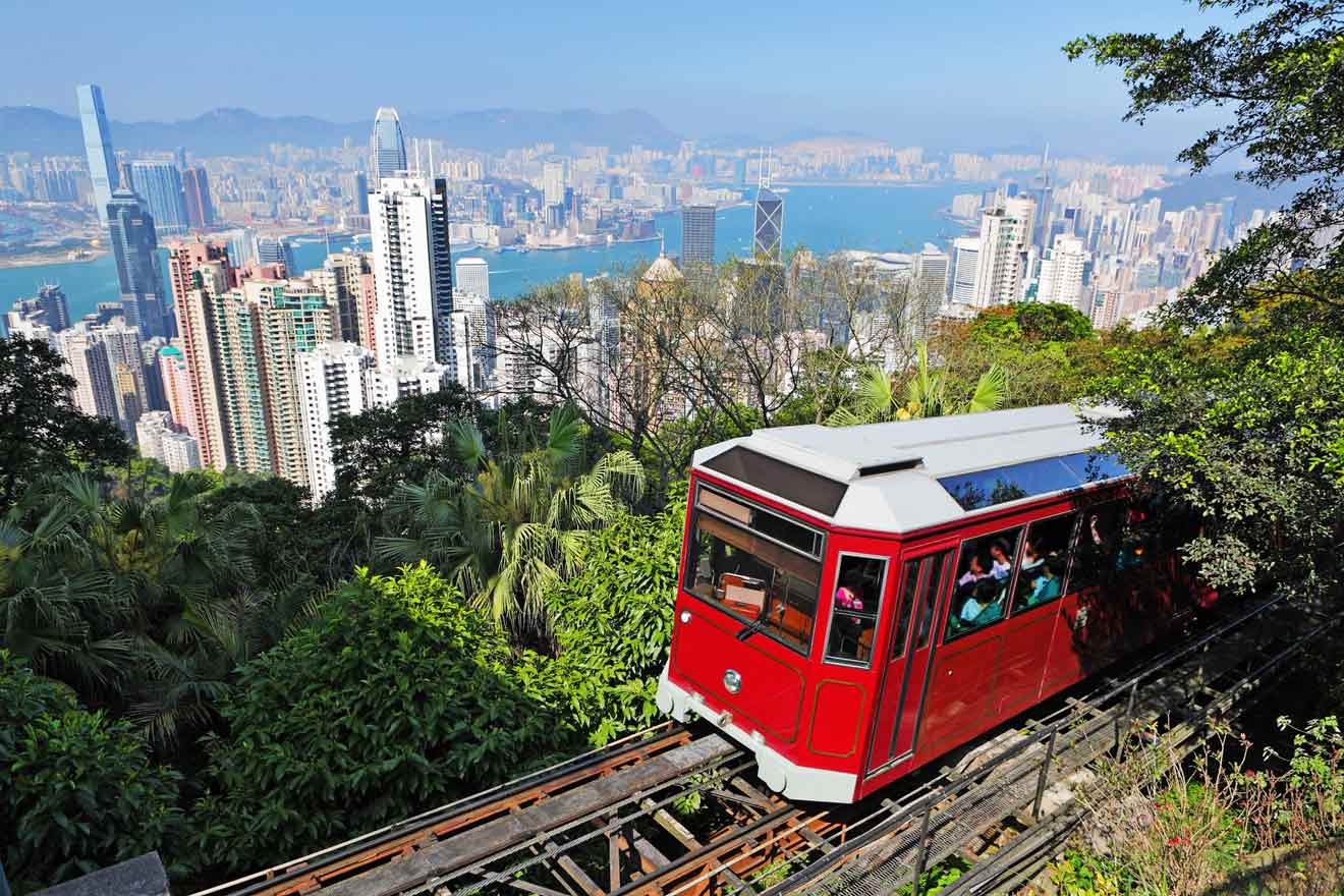 A red Victoria Peak tram ascending on the tracks with a stunning backdrop of the Hong Kong skyline and harbor, surrounded by lush greenery.
