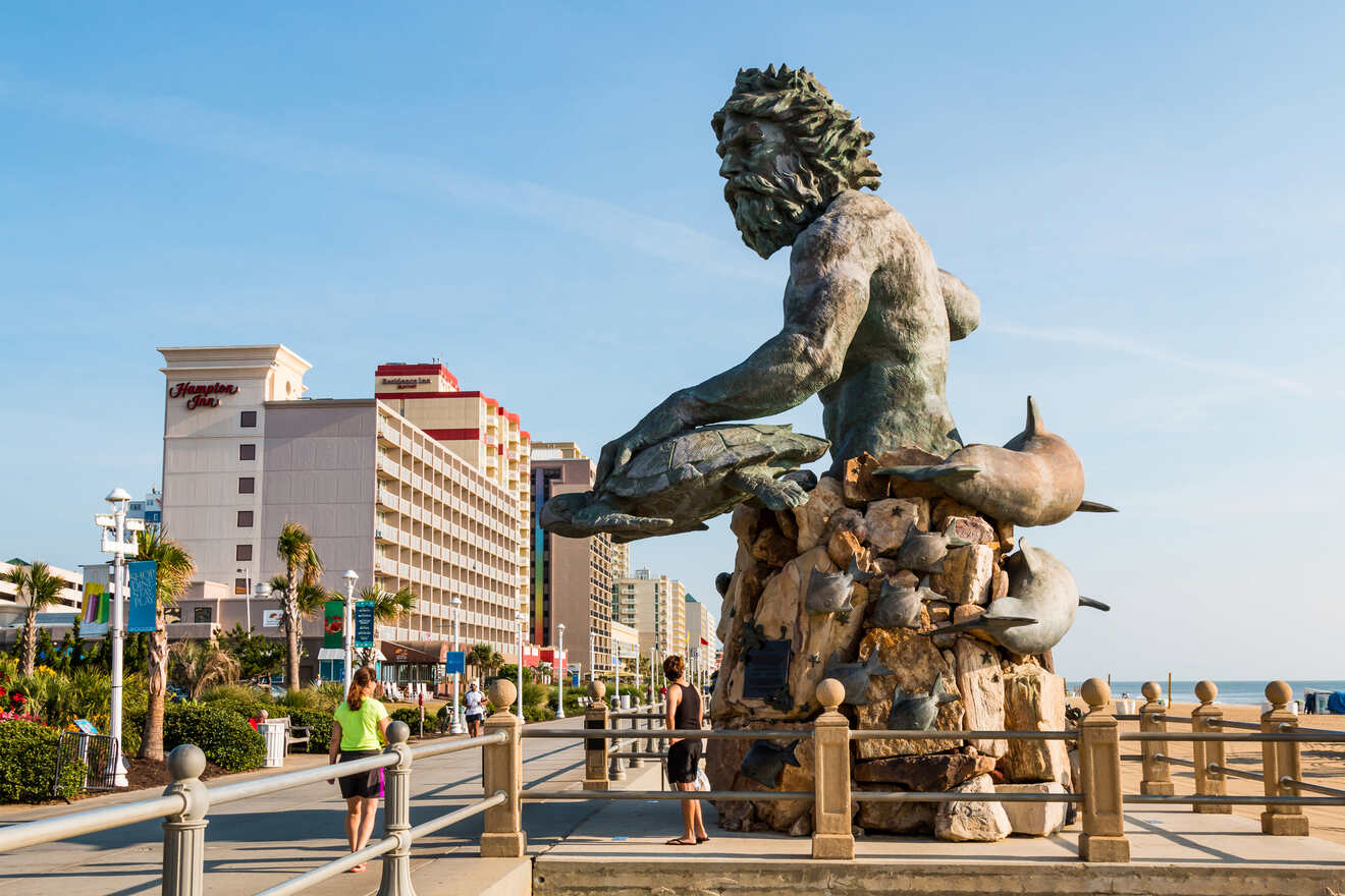 A striking statue of Neptune, a symbol of Virginia Beach, towering over the boardwalk with hotels in the background, under a clear sky