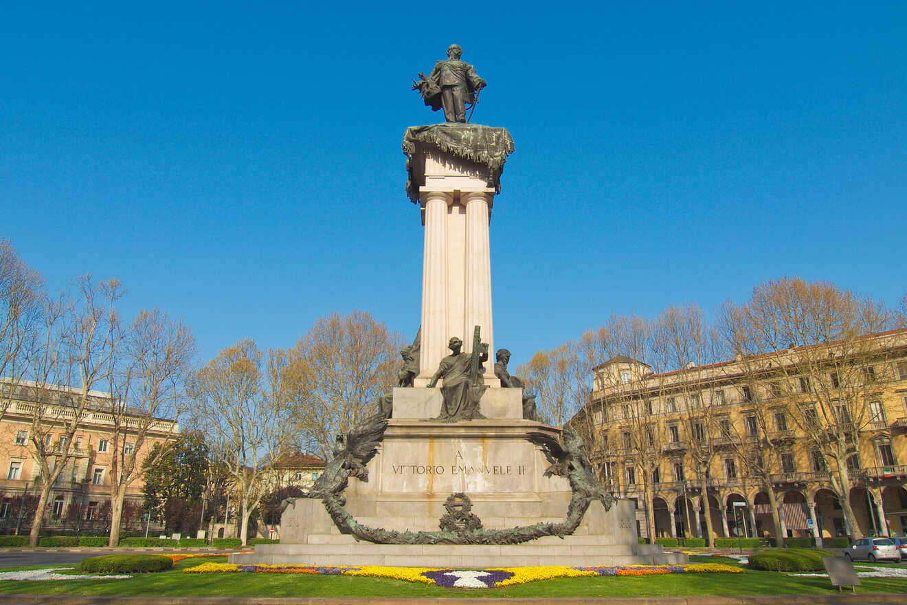 Monument to Vittorio Emanuele II in a square in Turin, Italy, flanked by statues and vibrant floral arrangements under a clear blue sky.
