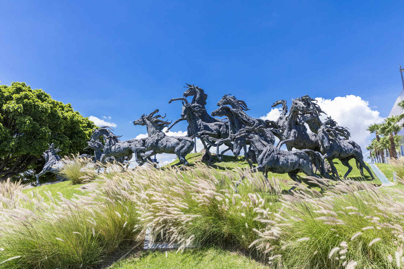 Dramatic sculpture of galloping horses and riders, captured in mid-motion, set against lush greenery and a bright blue sky