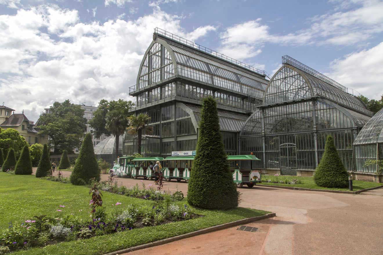 The Parc de la Tête d'Or greenhouse in Lyon, an imposing glass structure among well-manicured gardens with a tourist train passing by