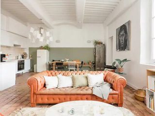 Spacious living room with a vintage leather sofa, white walls with decorative beams, and an open kitchen area, embodying a classic yet modern European style