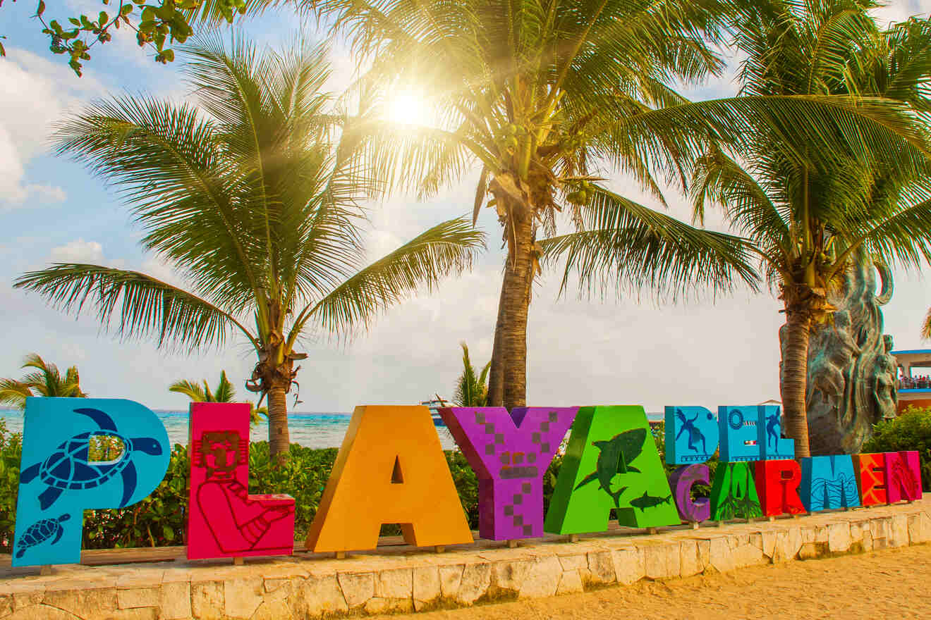 A vibrant sign spelling "Playa del Carmen" stands on a sandy beach with palm trees and the ocean in the background, under a bright sun.