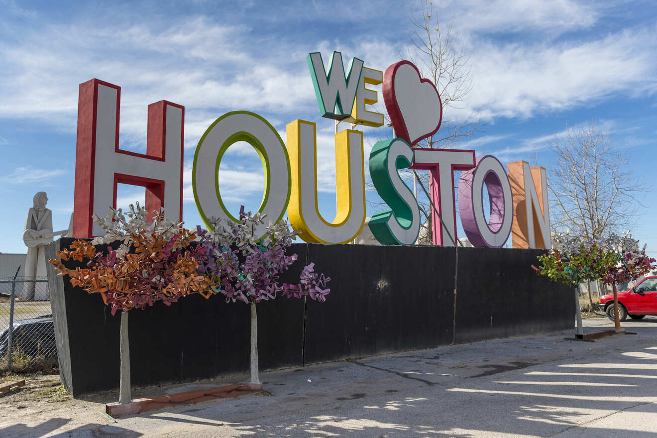 A large colorful sign reads "WE ♥ HOUSTON" with a red heart symbol in place of the word "love," situated outdoors against a blue sky backdrop.