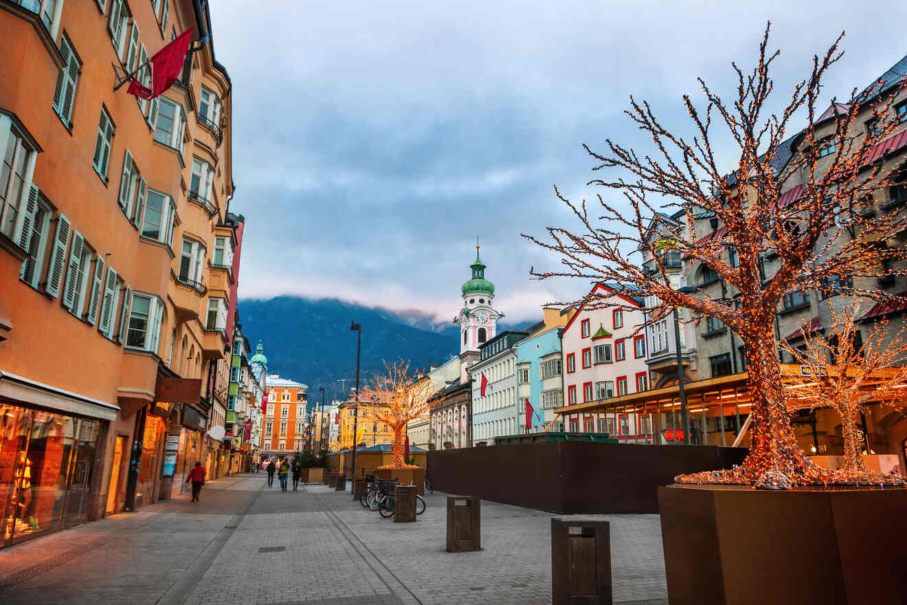 Twilight scene in an Innsbruck pedestrian zone with illuminated buildings, bare trees, and the Alps faintly visible