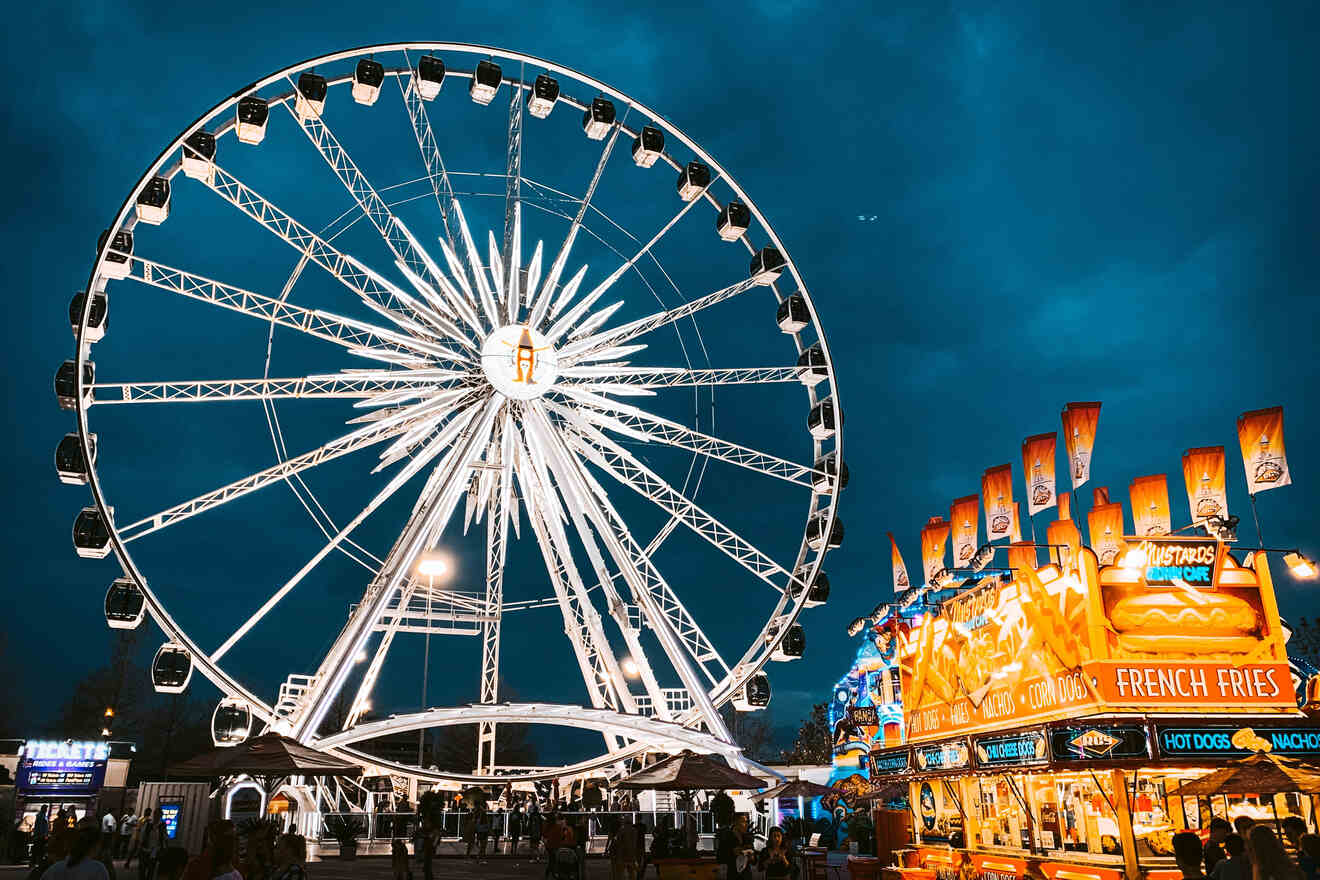 A large Ferris wheel illuminated against a dark evening sky with a brightly lit food stall offering French fries and hot dogs in the foreground.