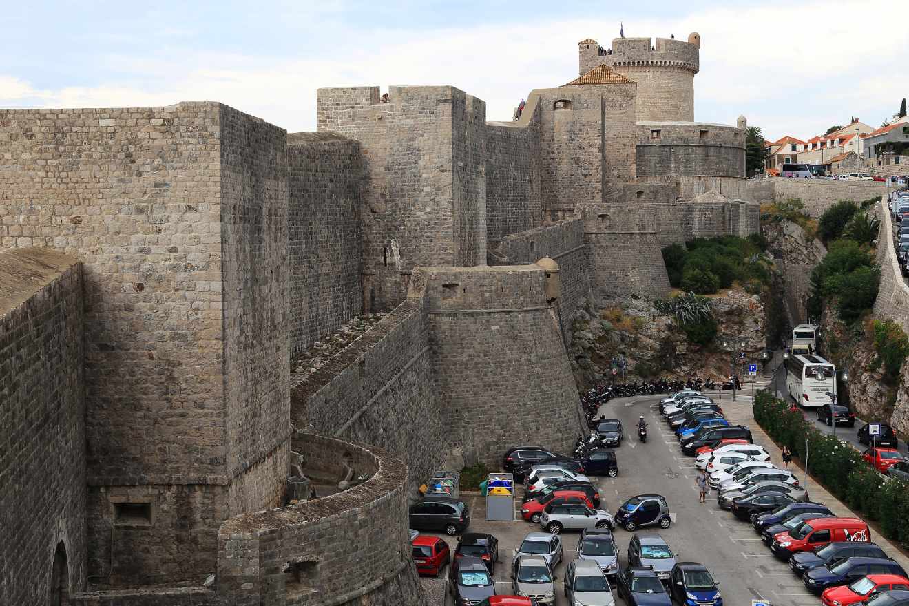 Massive stone walls of Dubrovnik's fortress, with cars parked along the road leading to the entrance