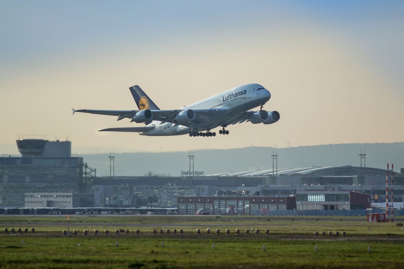 A Lufthansa Airbus A380 aircraft coming in for a landing at Bucharest Airport, with the terminal buildings in the background and a clear blue sky above