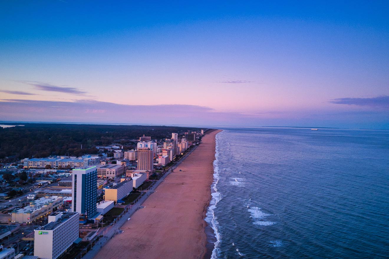 An aerial view of Virginia Beach at twilight, with the long stretch of sandy beach and ocean on one side and a line of hotels and buildings on the other.